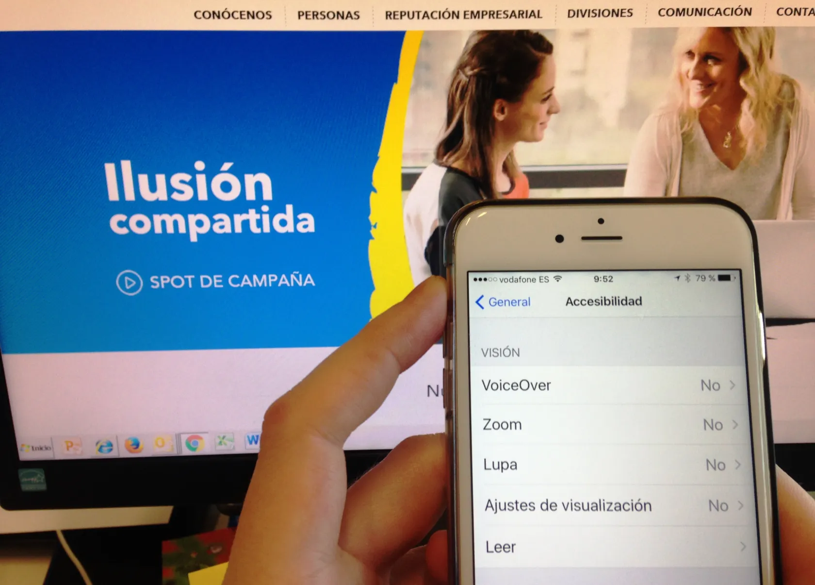 Se ve la pantalla de un teléfono en la sección de ajustes de accesibilidad y por detrás, en una pantalla de ordenador, la página web de ILUNION