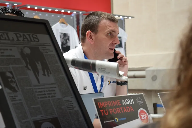 monitor con una portada de El País junto a un trabajador de ILUNION atendiendo a un visitante de un stand