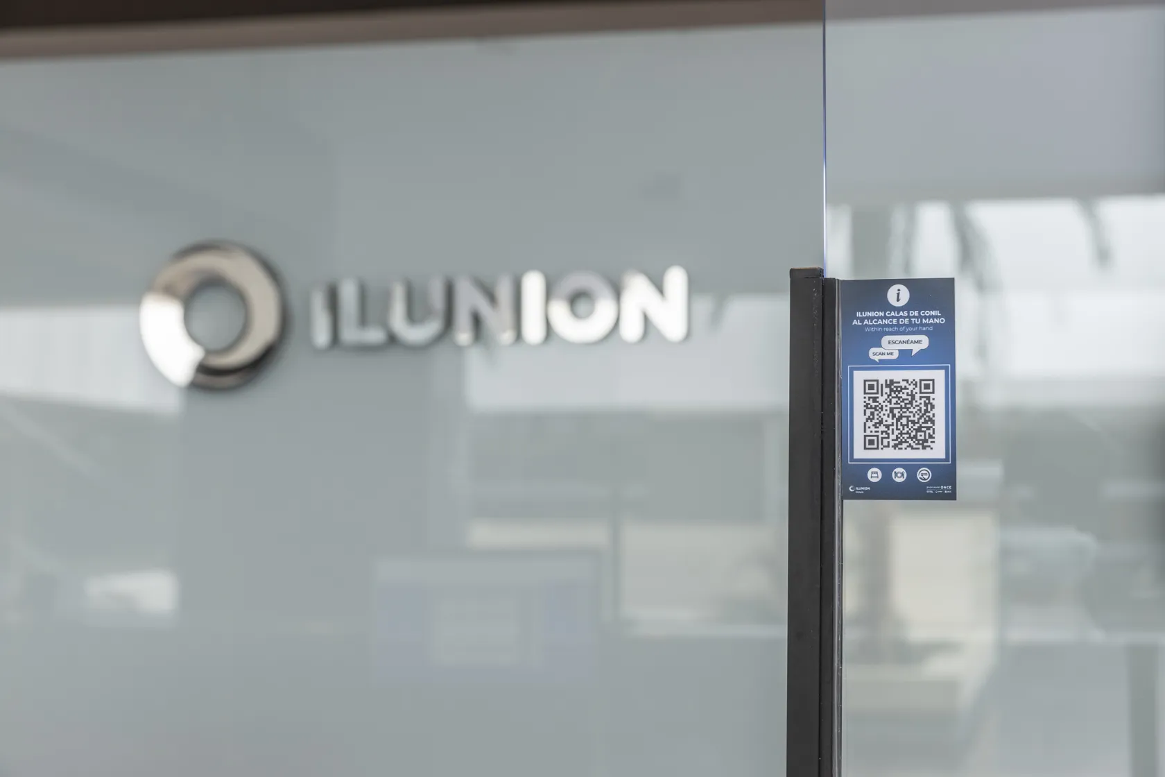Recepción de uno de los ILUNION Hotels. Al fondo, el logotipo del hotel, en primer plano una mampara de protección en la que hay una pegatina con un código QR de información