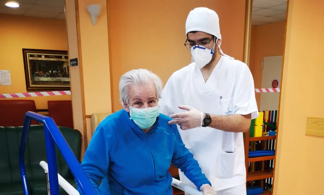 Una mujer mayor realiza ejercicio con una mascarilla y guantes, junto a un profesional, también con mascarilla y guantes.